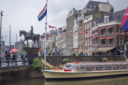 Amsterdam, Netherlands - 5th June, 2019: View of the city of Amsterdam from the canal to the bridge. Monument to Queen Wilhelmina surrounded by crowds of tourists walking under umbrellas on a cloudy summer day.のeditorial素材