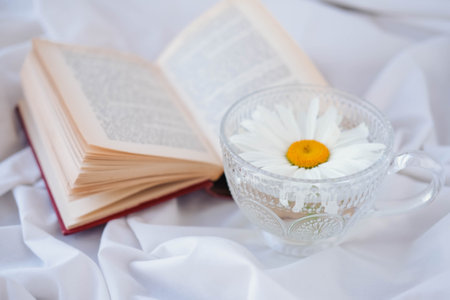 Open book with chamomile flower and cup of tea on white fabricの写真素材