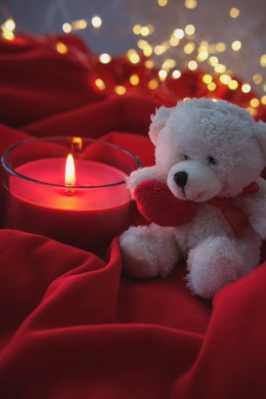 White teddy bear and burning candle on red fabric with bokeh backgroundの写真素材