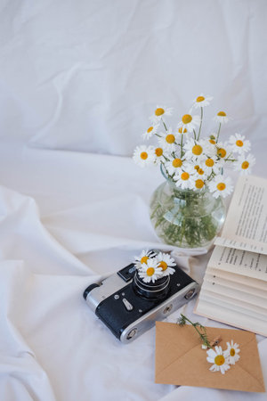 Bouquet of daisies in a glass vase, books and old camera on the bed