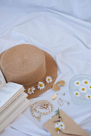 Straw hat, book, chamomile flowers, necklace and earrings on white bedding.の写真素材