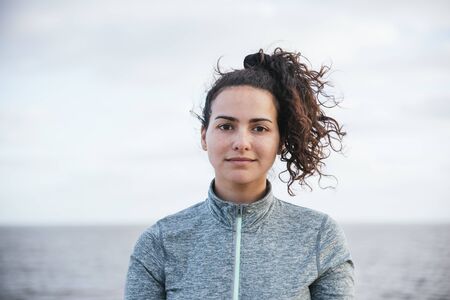 young latin girl looking at camera with the hair flying to the wind, with sportswear, smiling with the sea at backgroundの写真素材