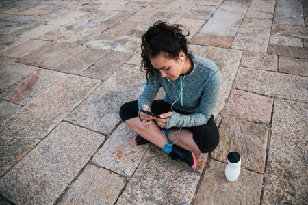 athletic girl sitting on the floor with her cellphone with headphones, and her bottle of water,...