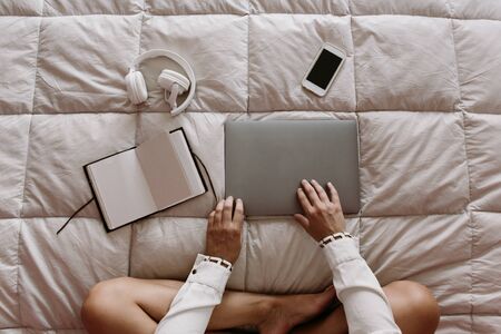 white, latin, caucasian girl sitting in bed, opening her computer. freelancer works at home. with mobile phone, laptop, notebook, notepad, draw tablet, external disk and headphones on the bed.の写真素材