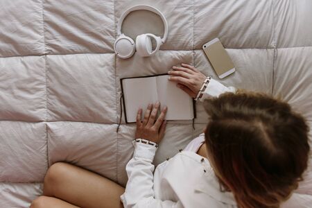 white, latin, caucasian, blond girl sitting in bed. freelancer works at home. with mobile phone, notepad and headphones on the bed.の写真素材