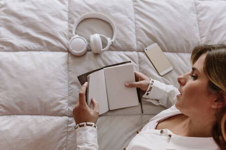 white, latin, caucasian, blond girl sitting in bed. freelancer works at home. with mobile, notepad, phone and headphones on the bed.の写真素材