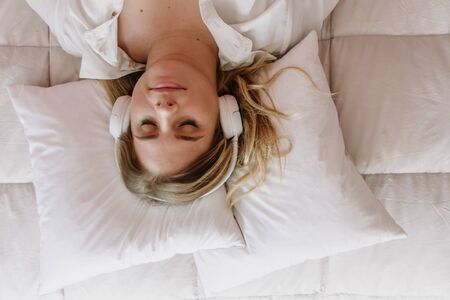 white, caucasian and blond girl, listening sleepy music, with white headphones, on the bed with cushions and a white padded, and a cellphone, computer, notebook.の写真素材