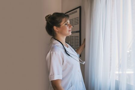 woman medical doctor looking through the window, with stethoscope around her neck and white jacketの写真素材