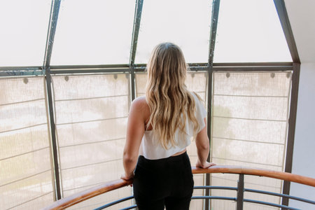 Caucasian, blond, young woman with her back to the camera, backwards, looking towards the window, with formal white shirt, and black pants.の写真素材