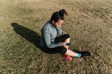 Female athlete resting and relaxing with mobile phone after workout, sitting on the grass. Healthy...