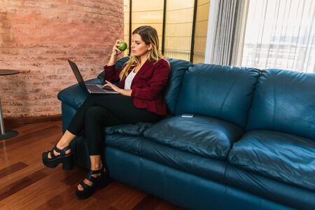 Stock photo of a caucasian woman sitting on her blue couch using her laptop. She is eating a green apple.の写真素材