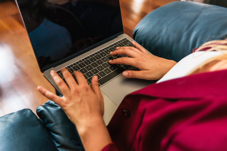 Stock photo of a caucasian woman sitting on her blue couch and typing on her laptop. She is...