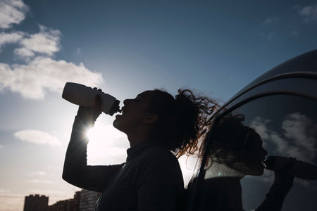 Stock photo of a woman leaning on a car drinking water. She is wearing sport shoes. She is...
