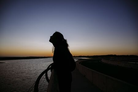 Stock photo of a caucasian woman looking to the side in the sunset. She is standing at a fence. She is unrecognizable.の写真素材