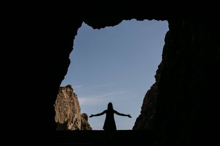 Stock photo of a silhouette of a woman standing in a cave entrance with her arms open. She is unrecognizable.の写真素材