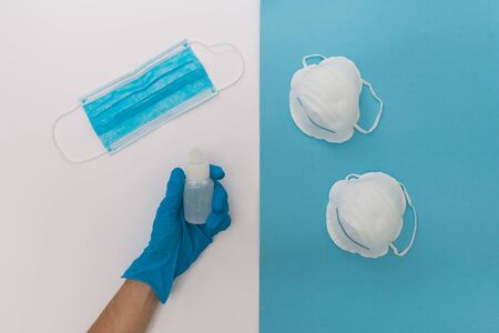 Stock photo of hands with blue medical latex gloves grabbing a bottle of sanitizer and medical mask on a blue and white background. The person is unrecognizable.の写真素材