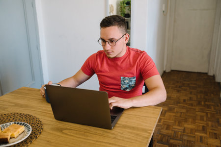 Stock photo of a caucasian adult man sitting at a table in front of his laptop and holding his head....