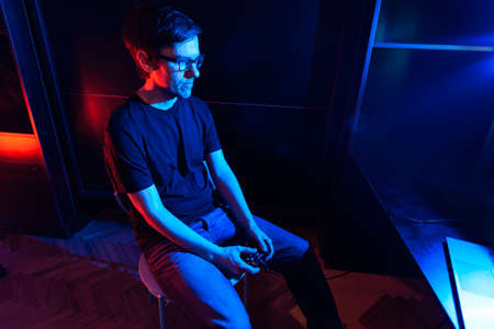Happy adult man sitting in his room and playing video games in his computer. There are red and blue lights in the room.の写真素材