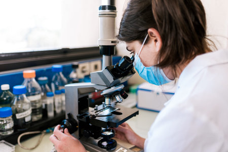 Stock photo of female scientist wearing face mask using a microscopy in her lab.