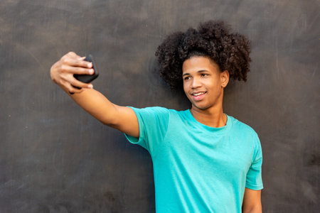 Afro latin young man taking selfie with mobile phone against a wall