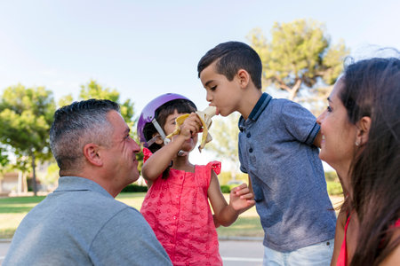 Two kids eating banana with parents sitting outdoors on the grass in a park.の写真素材