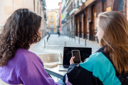 Two people using laptop and mobile phone while sitting outdoors at a coffee shopの写真素材