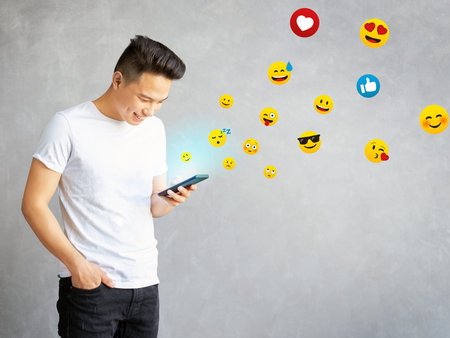 asian guy typing emoji on cell phone, young man having fun using smart phone isolated on gray...