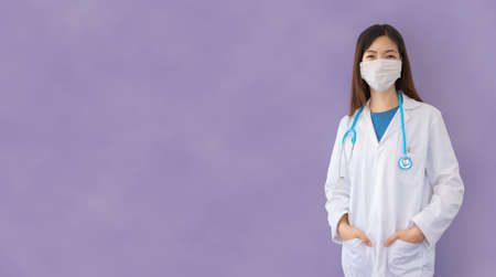 Young asian female doctor with lab coat and stethoscope protects herself with mask during coronavirus pandemic, smiling looks to the future with confidenceの写真素材