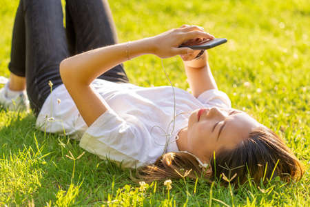 young girl lying on the grass listens to music texting in a park with sunset lightの写真素材