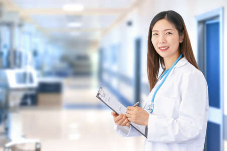 Smiling Asian female doctor wearing lab coat and stethoscope as she writes a document in her folder. In the blurred background you can see the hospital ward with medical equipment and patient roomsの写真素材