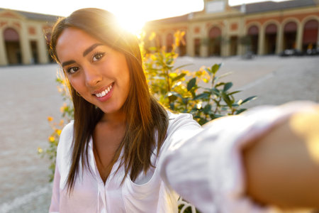 Young happy woman in white shirt making selfie standing in city square at sunset.