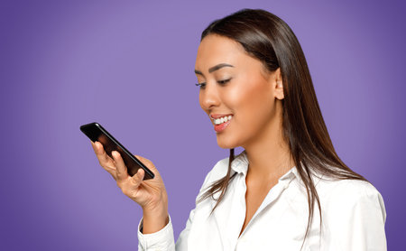 Headshot portrait of smiling young woman wearing white shirt and looking at her mobile phone...