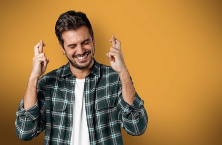Hopeful smiling young man keeps fingers crossed and eyes closed isolated on yellow colored...