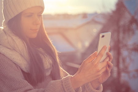 Beautiful young woman wearing warm knit clothes and using mobile phone in sunlight.の写真素材