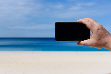 Men's hand holding blank screen smartphone or mobile on a beach. Vacation, travel concept.の写真素材
