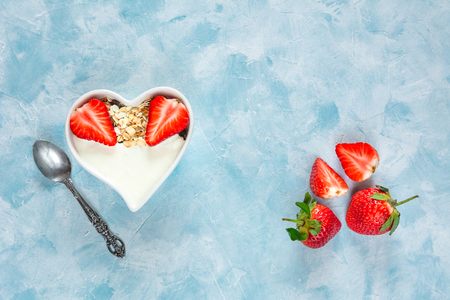 Yogurt with muesli and strawberries in a heart-shaped bowl on a pastel blue background.の写真素材