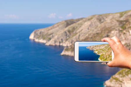 Hand holding smartphone and taking photo of beautiful cliffs of Porto Schiza on Zakynthos island. Greece.の写真素材