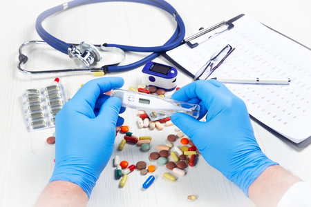 Doctor's hands in blue gloves holding thermometer and pills on the table