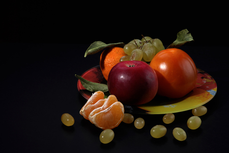 Tangerine slices and green grapes in front of a plate with fruit on a black background, the space under the textの写真素材