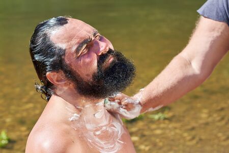 Shaving an elderly man with a black beard outdoors against the background of waterの写真素材