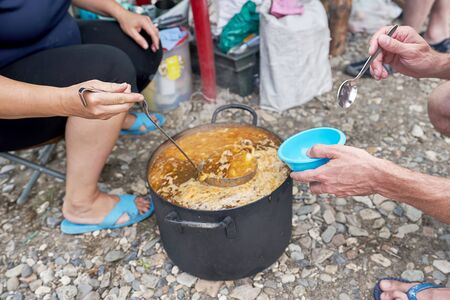 Distribution of food on a hike, a hand with a bowl and a spoon reaches for a large saucepan with soupの写真素材