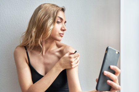 Beautiful young blonde with loose hair takes a selfie on a mobile phone and lowers the strap on her bare shoulderの写真素材