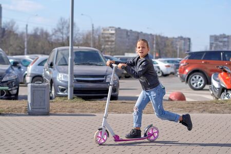 A nine year old girl in a leather jacket and jeans rides a pink scooter down the streetの写真素材