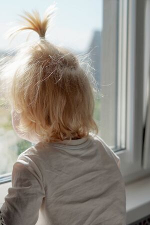A two-year-old blond child with a funny haircut on his head looks out the window. Frame from the back, sunlightの写真素材