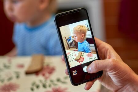 A hand holds a smartphone photographing a little blond boy in the kitchen sitting at a table with a lying piece of breadの写真素材