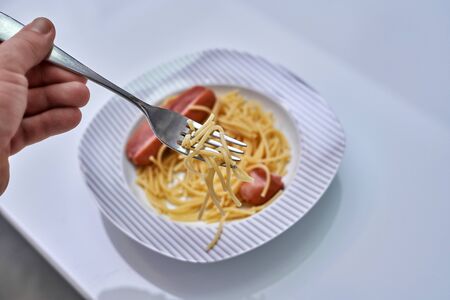 Hand with a spaghetti villa over a white plate with sausages and pasta, affordable foodの写真素材