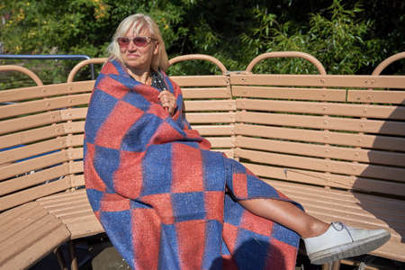 Blonde middle-aged wrapped a plaid blanket sitting on a wooden bench in Sunny dayの写真素材