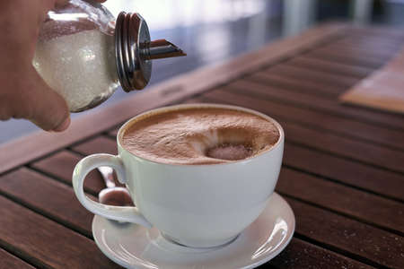 A hand with a sugar bowl pours sugar into the coffee. A white cup with a saucer stands on a wooden table.の写真素材