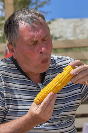Portrait of a middle-aged man eating a yellow ear of boiled corn. Vertical shotの写真素材