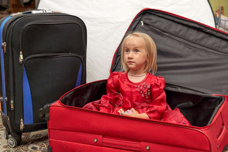 A girl with blond hair in a red dress sits in a red suitcaseの写真素材